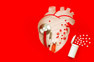 Stethoscope, pills, syringe on a electrocardiogram background, inside paper made heart on red backdrop