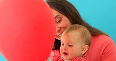 Happy young brunette mother and cute baby playing with red balloon on blue background
