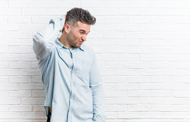 Young handsome man against a bricks wall touching back of head, thinking and making a choice.