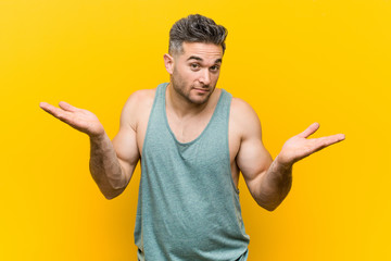 Young fitness man against a yellow background doubting and shrugging shoulders in questioning...