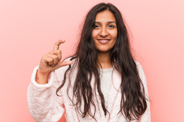 Young indian woman wearing pajama holding something little with forefingers, smiling and confident.
