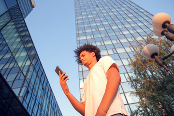 Fototapeta premium happy smiling urban hipster young man using smart phone. African american teenager holding mobile smartphone on sunset with lens flare
