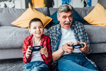 Active inspired man and his grandson enjoying new video game.. Sit on floor in living room.