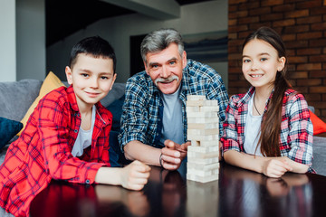 Happy cute little team playing game with grandpa, building constructor tower from multicolored wooden blocks..