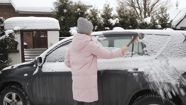 Young happy blonde woman in pink parka grey knit hat cleaning big black suv car from snow and ice, holding scraper brush tool in front of her house. Safe transportation winter driving concept.