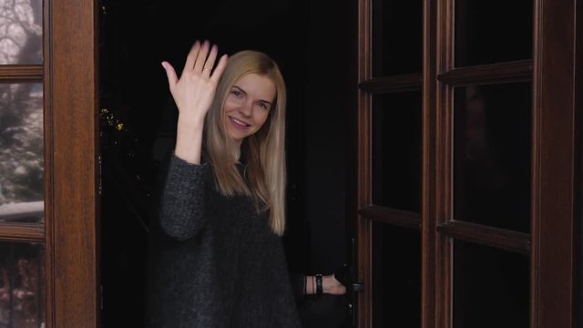 Young beautiful blonde woman with adorable smile dressed in grey sweater opens front door with snow winter yard reflections and welcomes a visitor, smiling, looking around and and invites to enter