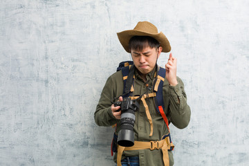 Fototapeta premium Young chinese explorer man holding a camera crossing fingers for having luck