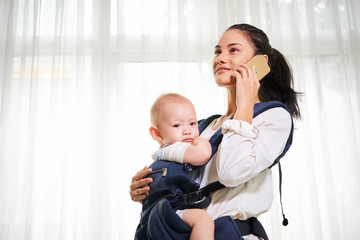 Positive young mixed-race woman carrying baby in sling when walking in house and talking on phone