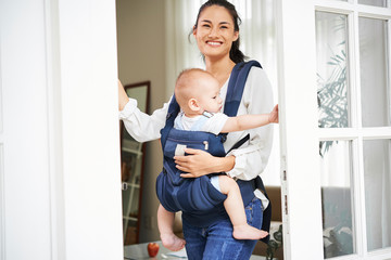 Happy young beautiful mixed-race woman carrying baby boy in sling when opening front door of her...