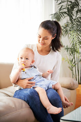 Smiling young woman sitting on sofa with baby chewing slice of tasty ripe apple