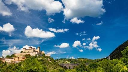 Fototapeta na wymiar View of Spoleto, green mountains, blue sky with white clouds. The Rocca Albornoziana fortress illuminated by the sun in summer. The bridge of the towers, Roman aqueduct. Trees in the foreground