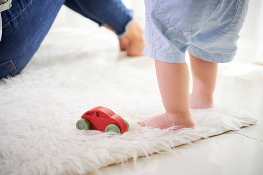 Little baby standing on fur carpet next to wooden car toy and sitting mother