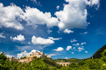 Fototapeta na wymiar View of Spoleto, green mountains, blue sky with white clouds. The Rocca Albornoziana fortress illuminated by the sun in summer. The bridge of the towers, Roman aqueduct. Trees in the foreground