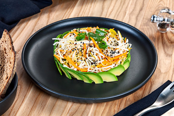 Close up view on vegan salad served on black plate on wooden background. RAW salad with carrot, cabbage, avocado. Healthy food concept. Copy space. Green vegan meal in bowl. Clean eating, dieting
