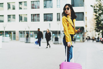 Side view photo of model look asian hipster female wearing sunglasses and modern outfit looking back while standing with a pink suitcase and a mobile phone in her hand on a blurred street background.