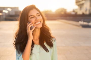 Happy female student talking on phone, walking at sunset in city