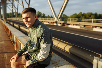 Young man in the sportwear sitting on a bridge and listening to music in the earphones