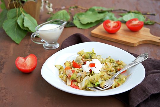 Fried cabbage with tomatoes in a white plate on a dark brown background. Served with sour cream.