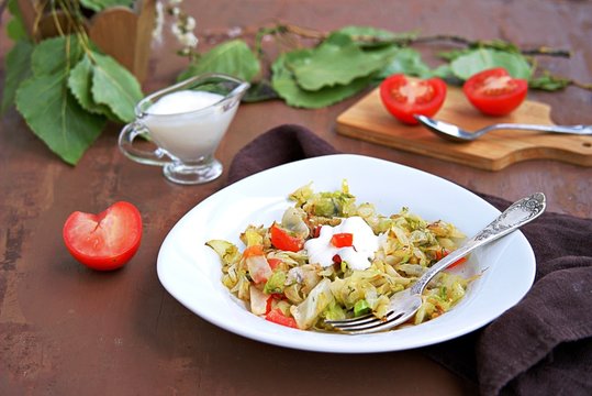 Fried cabbage with tomatoes in a white plate on a dark brown background. Served with sour cream.