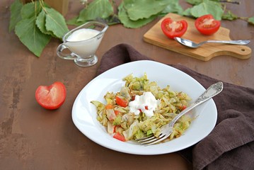 Fried cabbage with tomatoes in a white plate on a dark brown background. Served with sour cream.