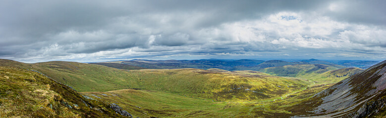 Fototapeta premium A panorama view of a Scottish mountain valley with heather and stream under a stormy huge white clouds sky