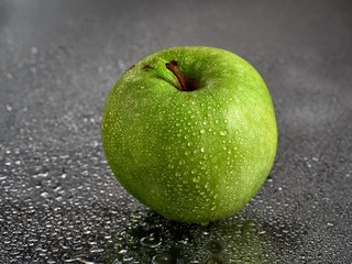 A ripe green Apple lies on a black wet glass and drops of water fall from it.Drops run down on the glass