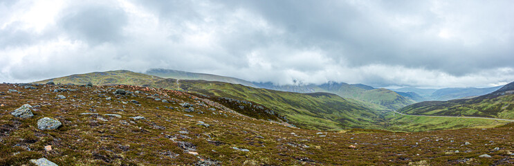 A panorama view of a Scottish mountain valley with a road and mountain range under a stormy huge white clouds sky