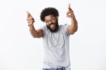 You rock man. Portrait of joyful good-looking emotive dark-skinned guy with afro hairstyle and beard pointing finger pistols at camera with raised hands and broad smile as liking dance moves of friend