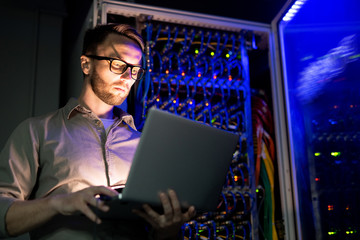 Serious busy young IT engineer in glasses standing against network server equipment with glowing...