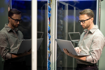 Serious busy young IT engineer with beard standing by server cabinet and using laptop while...
