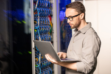 Serious thoughtful handsome young server engineer in glasses standing in database center and using...