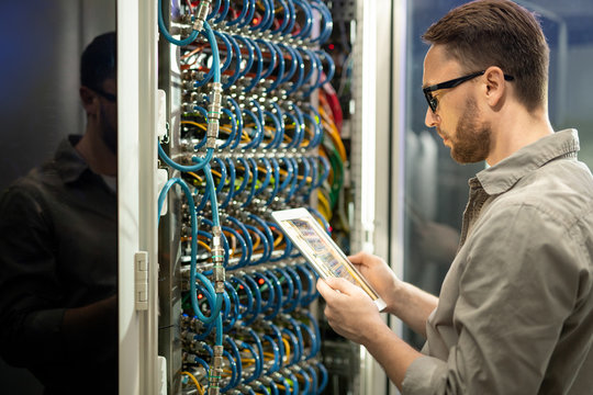 Serious pensive young data center technician in glasses standing against cabinet of server and using digital tablet while checking connections