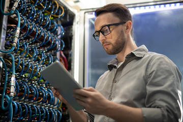 Serious smart young system engineer with beard standing in server room and using tablet while...