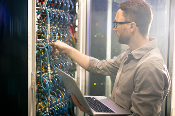 Serious busy young male engineer in glasses standing against mainframe and adjusting cables while...