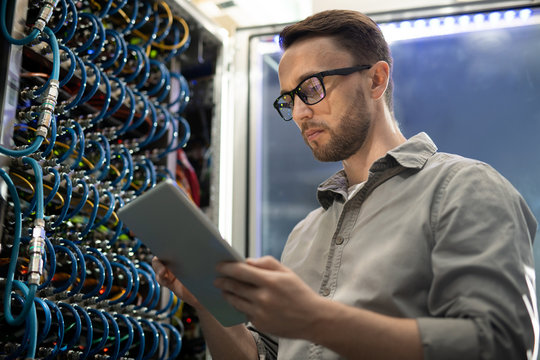 Serious smart young system engineer with beard standing in server room and using tablet while testing connectivity through app