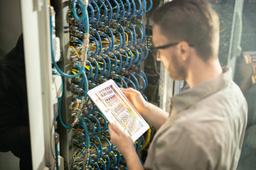 Busy server engineer standing by open cabinet of network server and using modern device while...