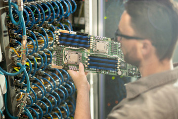 Busy skilled young IT expert in glasses standing by server cabinet and analyzing motherboard of...
