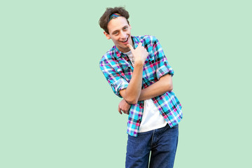 Portrait of cunning young man in casual blue checkered shirt and headband standing and looking at camera with cheating face and sly smile. indoor studio shot, isolated on light green background.