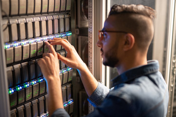 Serious busy hipster IT specialist standing by open cabinet and examining network racks while analyzing provider of server