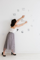 Young beautiful caucasian woman in a long skirt transfers big clock hands on a white wall