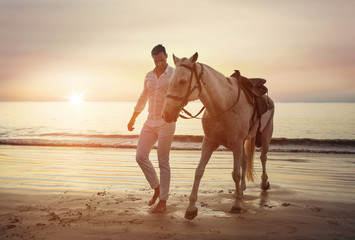 Handsome, young man walking with a stallion alongside the coast
