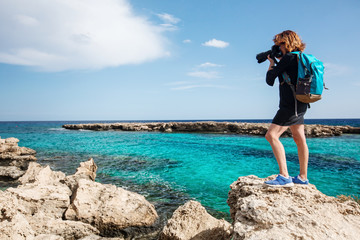 The young woman the traveler with a backpack and in a dress costs with the camera on the bank of the beautiful blue sea. It takes the picture.