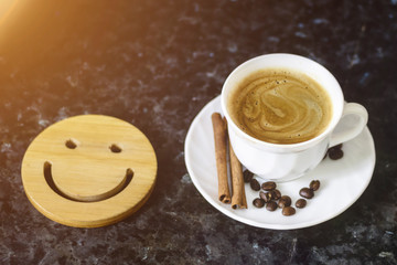 A cup of coffee is the key to a good mood. Wooden smiley on a dark, black, textural background. On the edge you can see a white cup with espresso, on the saucer are coffee beans and cinnamon sticks