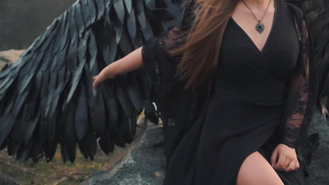 smooth panoramic shooting, girl with bright delicate hand gently smoothes large feathers of her strong wings, lady in long black dress with loose lace sleeves sits on stone near river, golden sunset