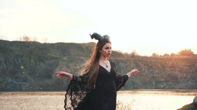 in a long vintage black dress with loose lace sleeves, a witch with big horns on her head raises her arms to the sides and enjoys wind during sunset