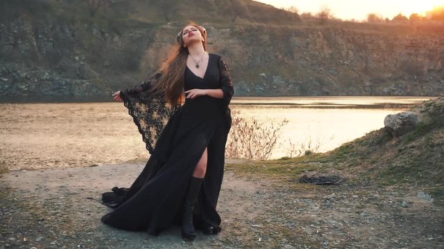 unusual awesome video of a mythical creature, a lady with a creative dark make-up, girl in long black vintage dress horns, Satan angel with green magic pendant, arms to sides and enjoys wind