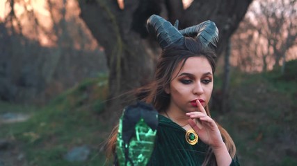 mysterious mythical creature with large sharp black horns on head holds finger at red bright lips, girl in long green emerald mantle with magic stick in her hands. witch asks to keep her secret - Powered by Adobe