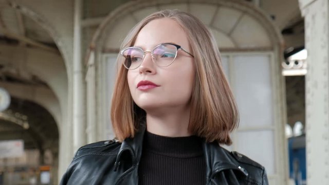 Attractive red-haired young woman with short haircut, wearing glasses and black leather jacket is standing at beautiful historic Vitebsk railway station in St. Petersburg and waiting for train.