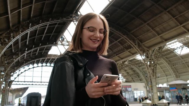 Attractive red-haired young woman with short haircut, wearing glasses and black leather jacket is standing on railway station in St. Petersburg and waiting for train. Uses mobile phone.