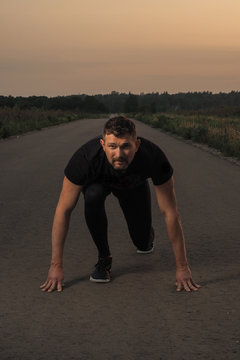 Mature fitness coach or runner in black sport joggers and tee stands in getting ready pose on a free countryside road in the evening time
